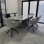 A modern dining set featuring a rectangular gray stone-top table with a sculptural ribbed pedestal base, surrounded by six light gray upholstered swivel chairs with black metal star bases, set in a contemporary room with light wood flooring and large black-framed windows.