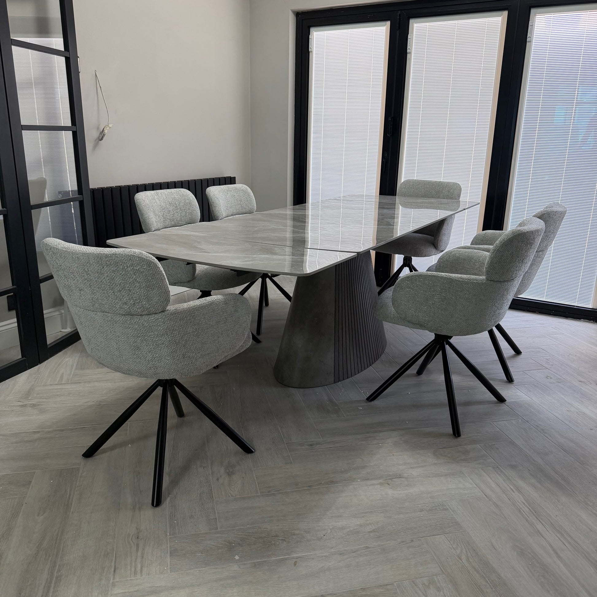 A modern dining set featuring a rectangular gray stone-top table with a sculptural ribbed pedestal base, surrounded by six light gray upholstered swivel chairs with black metal star bases, set in a contemporary room with light wood flooring and large black-framed windows.