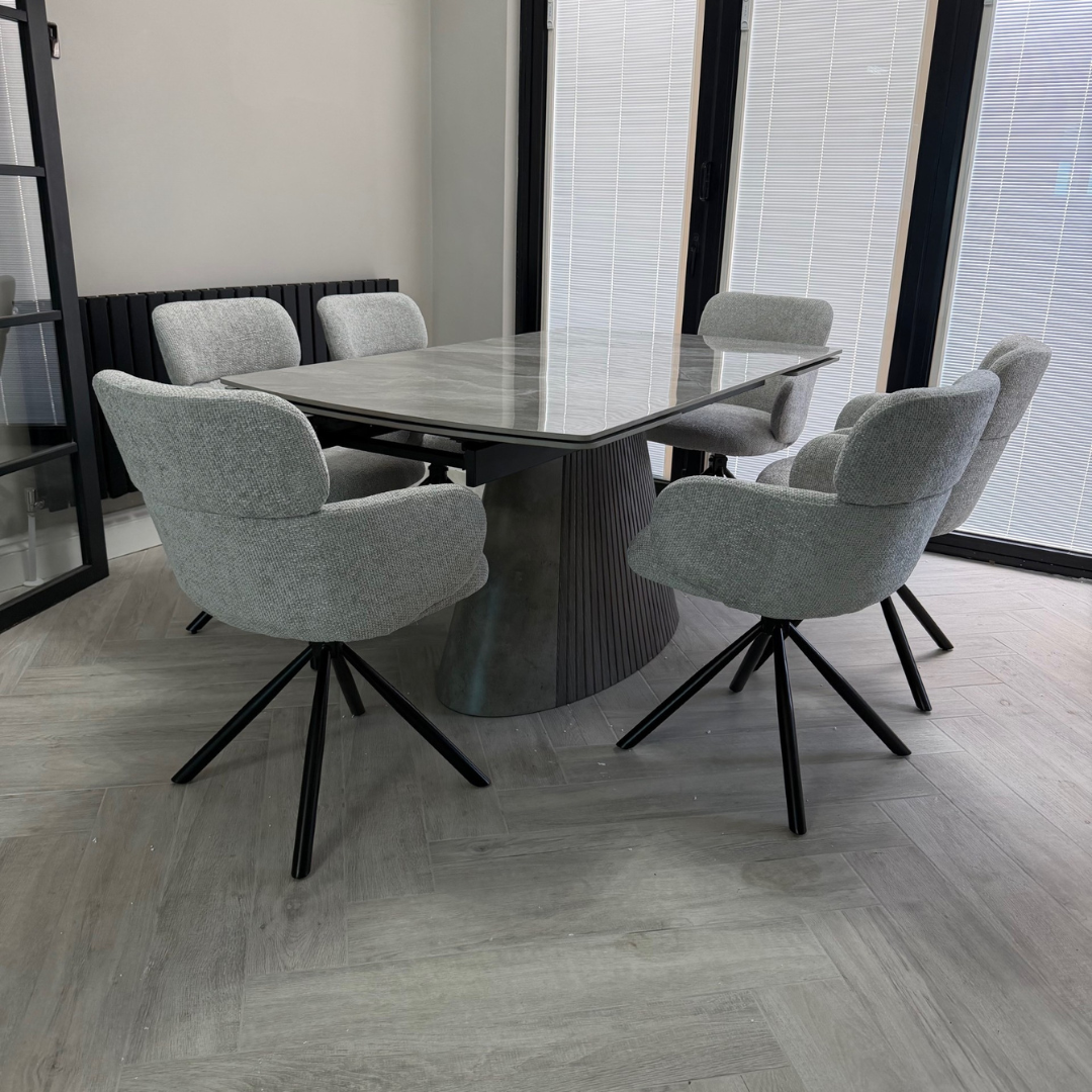 A modern 6-piece dining set featuring a rectangular gray stone-look table with a fluted pedestal base, surrounded by six light gray upholstered swivel dining chairs with black metal legs, set in a bright room with gray wood-look flooring.